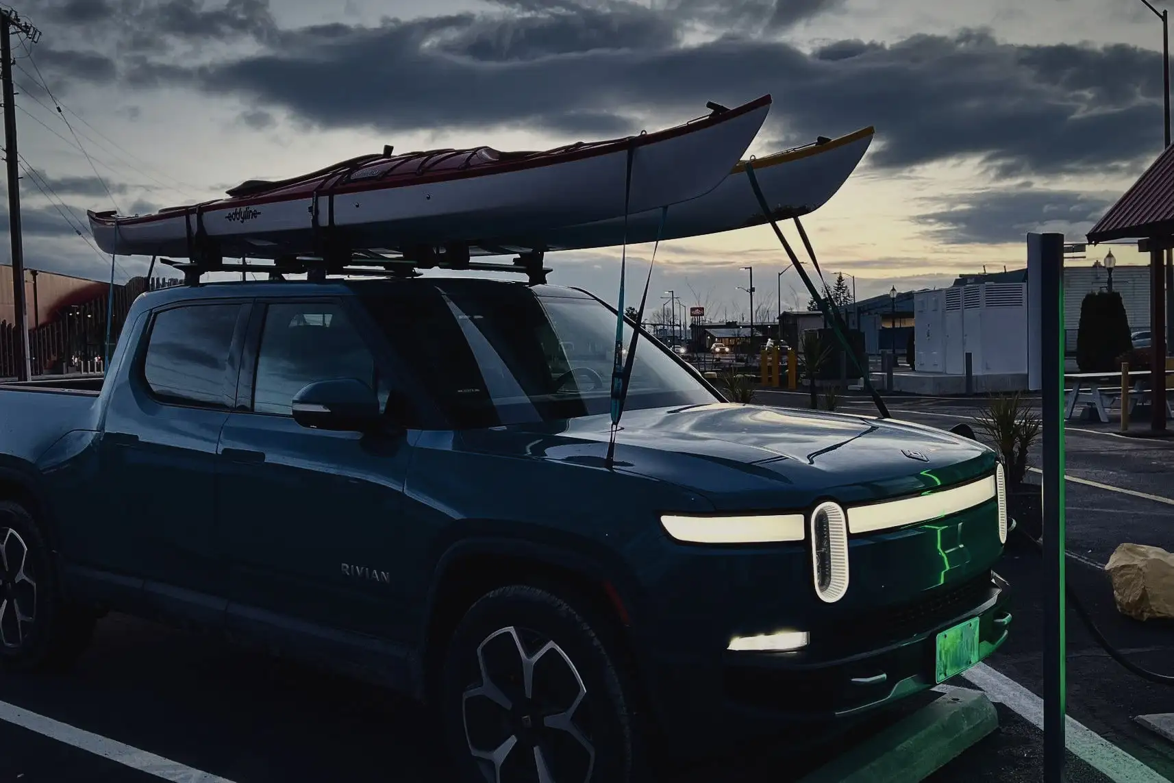 Rivian R1T at dusk with its front light bar lit, kayaks on the roof, under a storm-blue sky