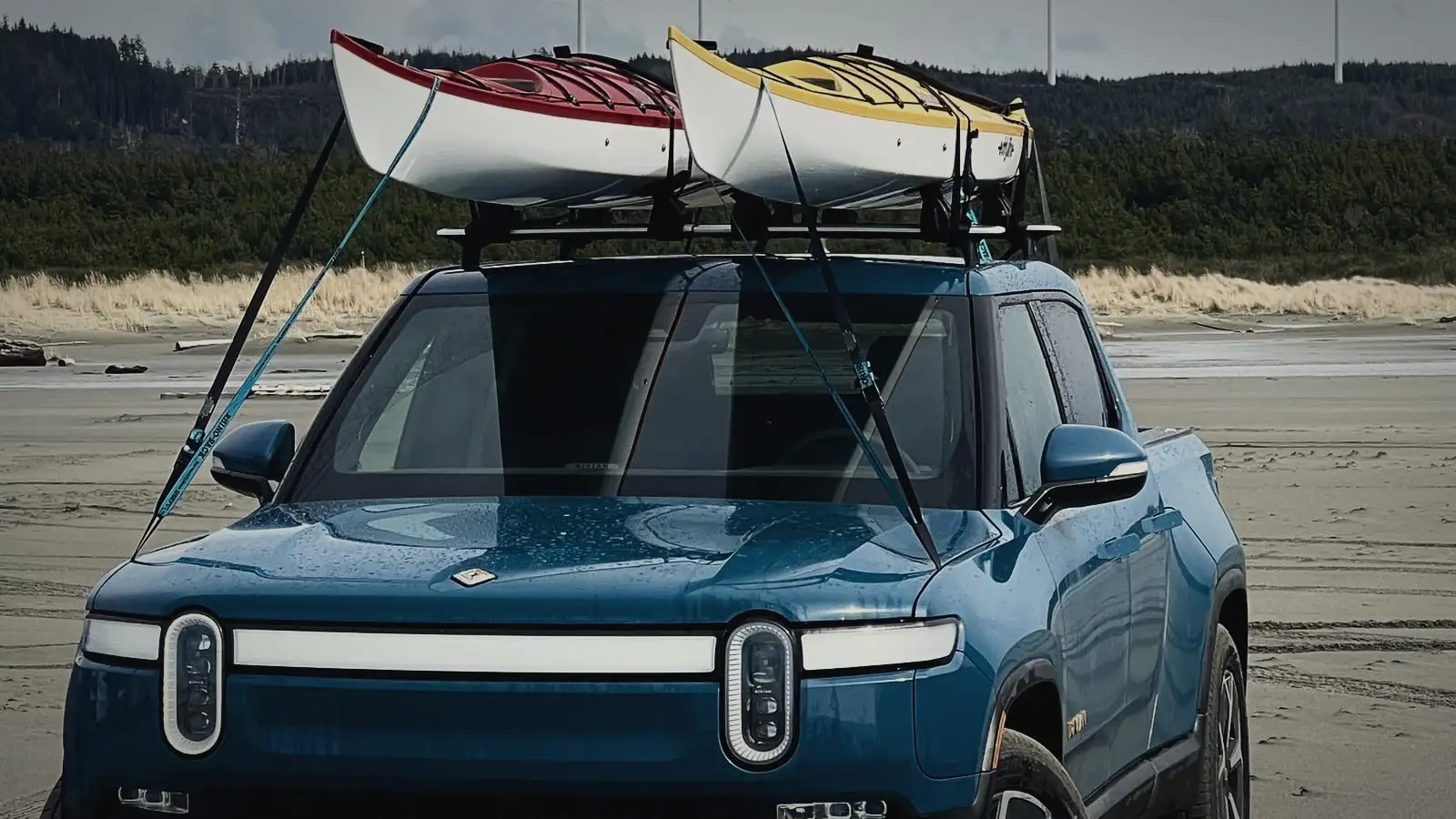 Rivian R1T head-on on a beach, light bar lit, two kayaks on the roof rack