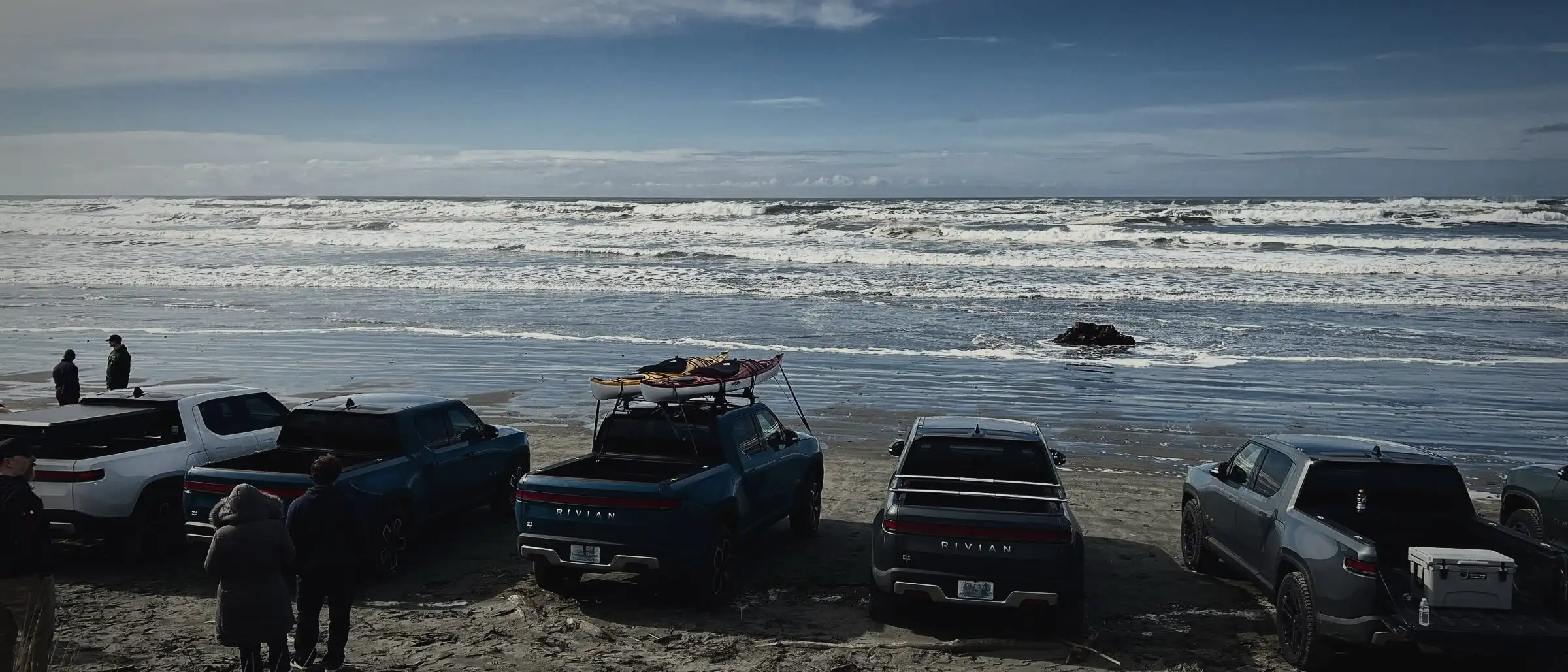 A line of Rivian trucks parked on the sand facing the surf under an overcast sky